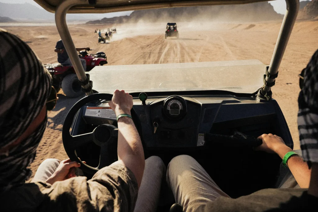 Vue depuis le cockpit d'un buggy en piste dans le désert, mains du conducteur sur le volant, convoi de quads et de buggys soulevant la poussière devant
